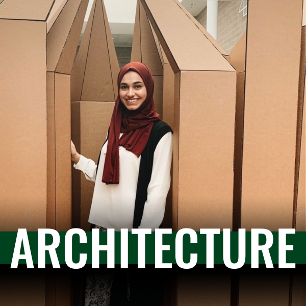 female student wearing a white shirt and a hijab standing inside a hand made cardboard structure